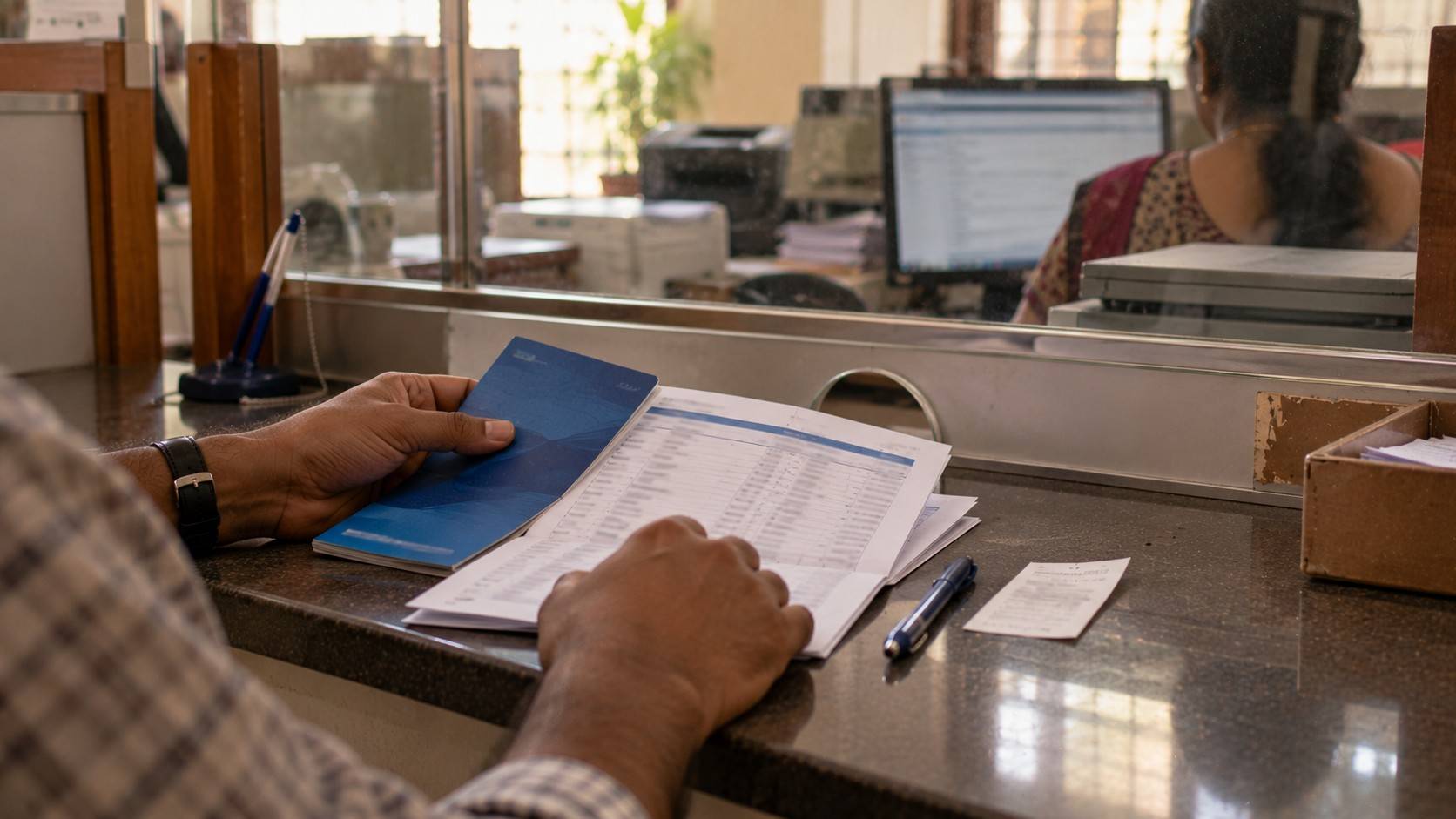 A person at a bank branch counter holding a passbook and a printed account statement